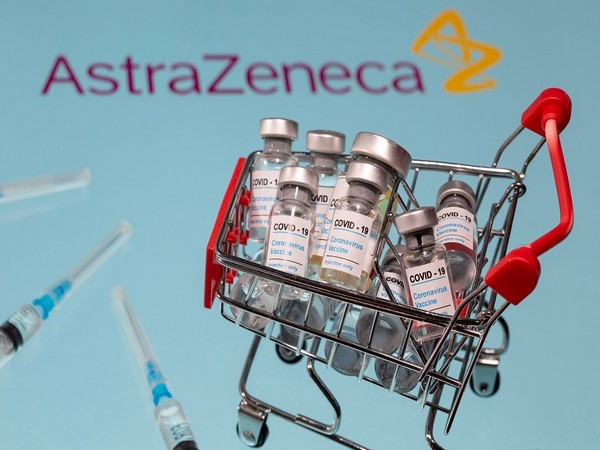 A small shopping basket filled with vials labeled "COVID-19 - Coronavirus Vaccine" and medical sryinges are placed on a AstraZeneca logo